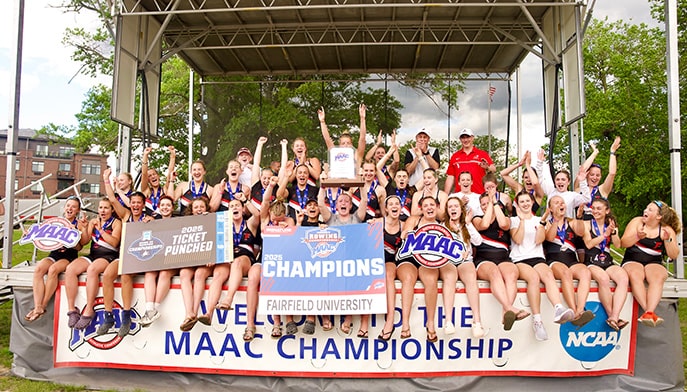 Fairfield's Women's Rowing team posing on a stage with their 2025 MAAC Championship winner banners, cutouts, and trophy.