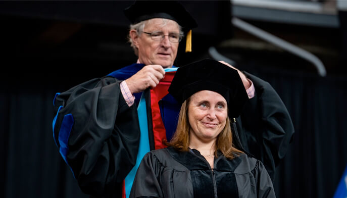 A woman in a graduation gown stands in front of a man wearing a graduation cap, both smiling proudly at their achievement.