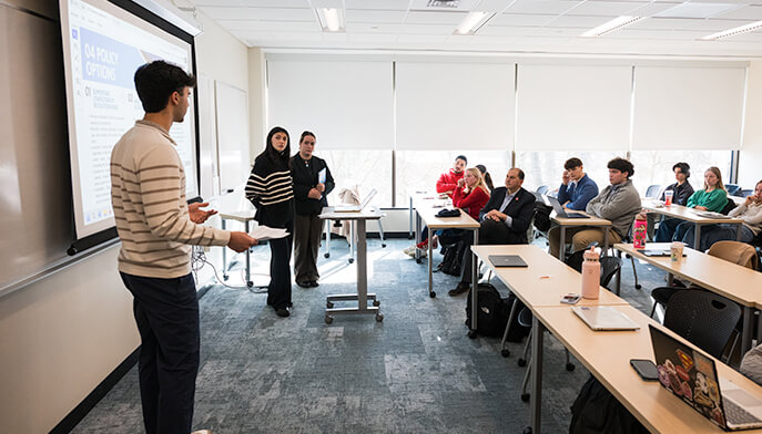 A male student presents to a classroom of engaged students, using a projector and visual aids to enhance his lecture.  