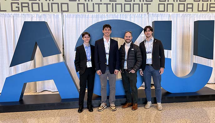 Four men pose and smile in front of large blue AGU letters at a conference or event, all wearing name badges and business casual attire, with a white curtain backdrop.