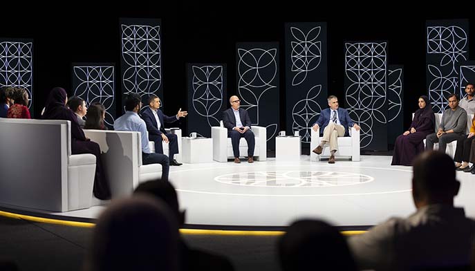 A group of people sit in a circular studio, engaged in a panel discussion. Three men sit on chairs in the center, while others sit on sofas around them. Decorative geometric panels are displayed in the background.