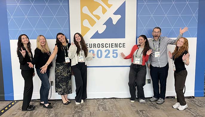Seven smiling people stand in front of a wall with a “Neuroscience 2025” sign, posing with cheerful and welcoming gestures. 