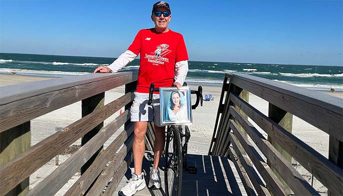 A man in a red shirt stands on a wooden boardwalk by the beach with a bicycle. Attached to the bike is a framed photo of a woman. Ocean waves and a clear blue sky are visible in the background.