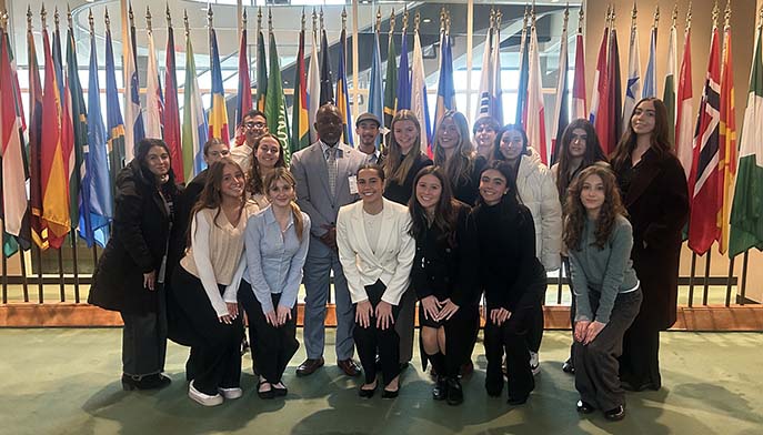 A diverse group of young adults and one man in a suit pose together in front of a display of various international flags inside a formal building.