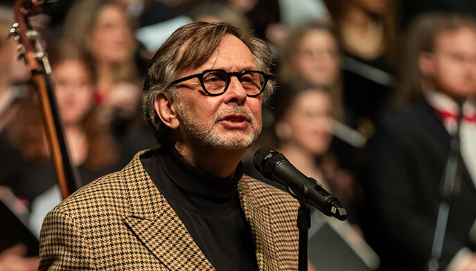 Fairfield University professor of music Brian Q. Torff stands wearing a plaid jacket and glasses, speaking into a microphone while holding a paper. A choir stands blurred in the background, creating a formal and musical atmosphere.