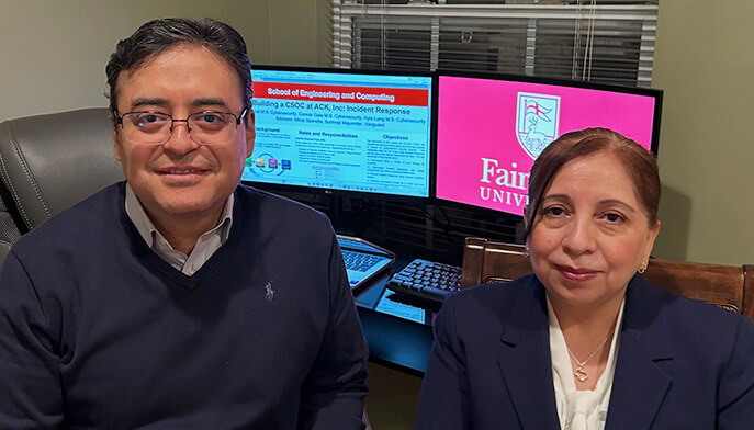 Two people are sitting in an office, smiling at the camera. Behind them, a computer monitor displays a presentation and a university logo. The atmosphere is professional and warm.