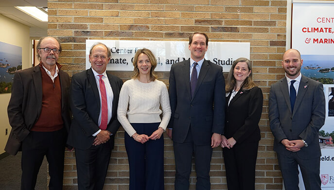 Group of six people posing in front of a brick wall with a sign related to climate studies. They are smiling, dressed in business attire, conveying a professional yet friendly atmosphere.