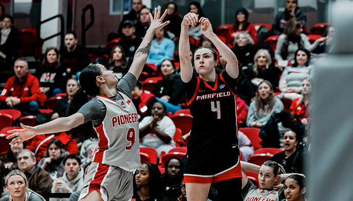 Kaety L’Amoreaux ‘27 in action wearing a black "Fairfield" jersey shooting, while another in a "Pioneers" jersey tries to block. The crowd watches intently.