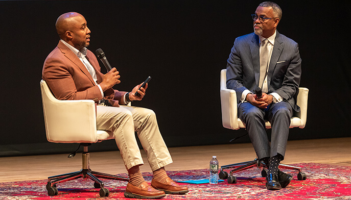 Don Sawyer III, PhD, and Eddie S. Glaude Jr., PhD, seated on white chairs on a stage with a red patterned rug. One speaks into a microphone, creating a focused, professional atmosphere.