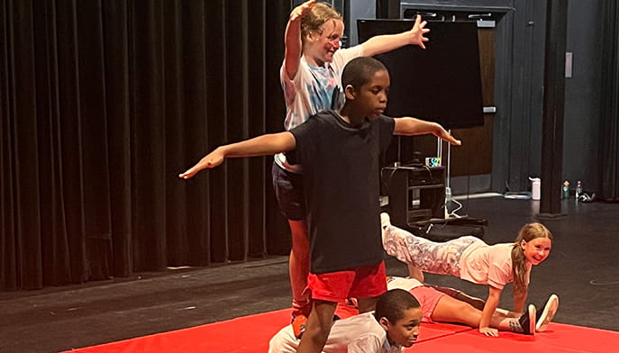 A group of local children building a human pyramid on a red gym mat as part of a circus workshop.