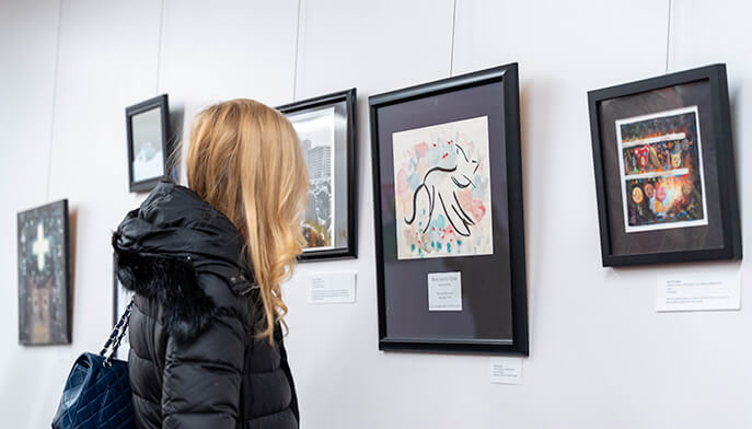 A woman with long blonde hair in a black coat admires framed abstract artwork in a gallery. The scene conveys a sense of curiosity and appreciation.