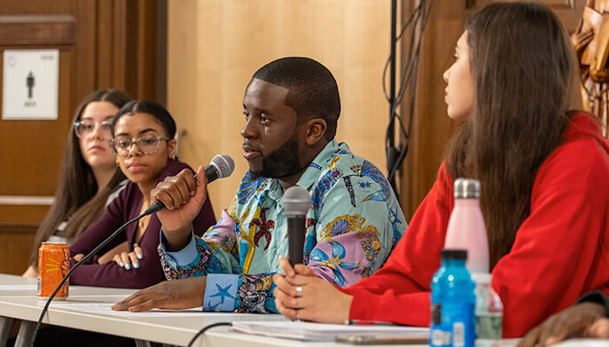 A diverse panel of people at a discussion table. A man in a colorful shirt speaks into a microphone, engaging the attentive audience.