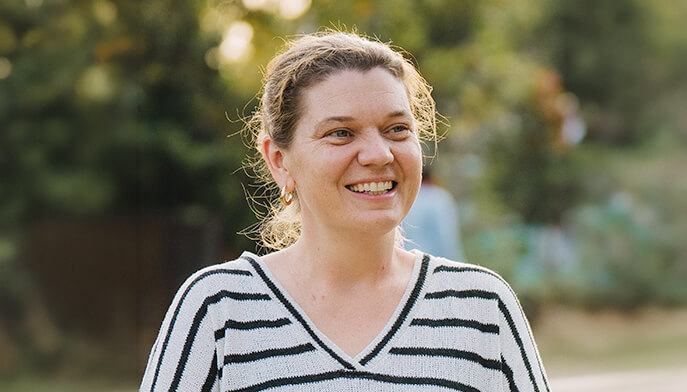 A smiling Maggie Doyne stands outdoors, wearing a white and black striped sweater. The background is a sunlit, blurred green park, conveying a joyful and serene mood.