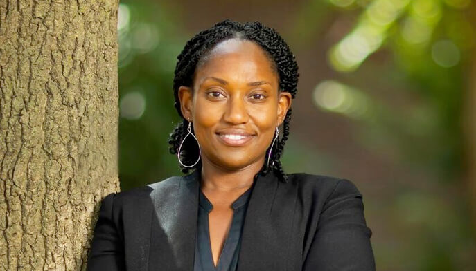 Marcia Chatelain with braided hair smiles confidently, standing beside a tree in a sunlit outdoor setting. She's wearing a black blazer and hoop earrings.
