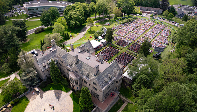 Aerial view of a Bellarmine Hall surrounded by lush trees. Nearby, a crowd gathers on a lawn for an outdoor event, suggesting a celebratory atmosphere.