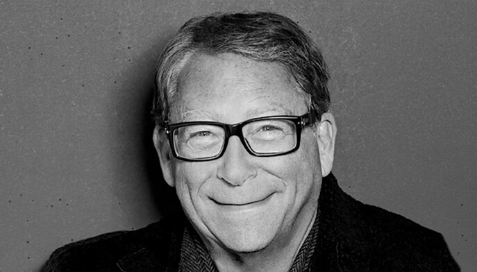 A smiling Stuart Weitzman with glasses stands against a solid gray background. The black-and-white image has a warm, approachable tone, conveying friendliness.