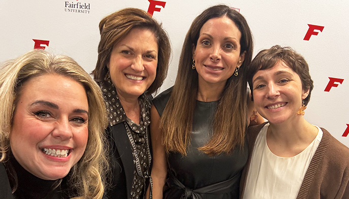 Four women smiling at the camera, standing closely together against a backdrop with "Fairfield University" and red "F" letter. Casual, friendly atmosphere.