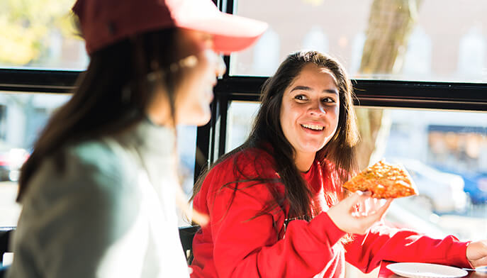 Two students at Colony Grill share a lively conversation. One, in a red sweatshirt, holds a slice of pizza, smiling warmly. The scene is bright and cheerful.