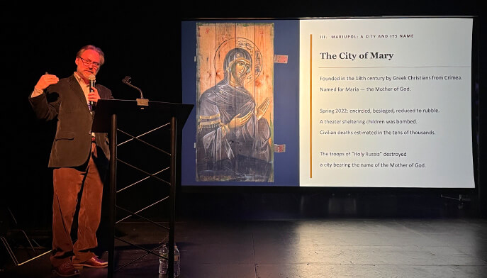 Sergei Chapnin at a podium speaks, gesturing, beside a screen displaying a religious icon and text about Mariupol.