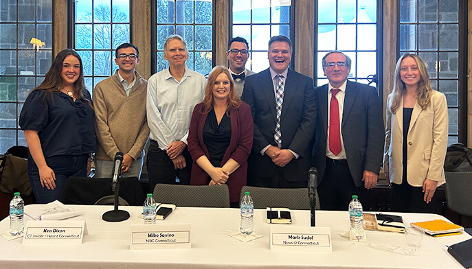 A group of eight people, dressed in business attire, pose smiling in a conference room. Behind them are large windows showing a tree outside.