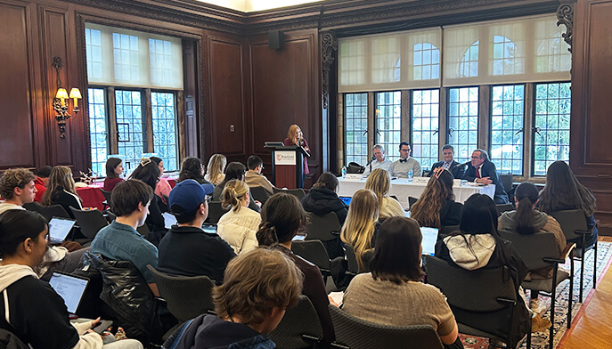 A panel discussion in a wood-paneled room, with people seated facing four speakers at a table. A woman stands at a podium, and the mood is attentive.