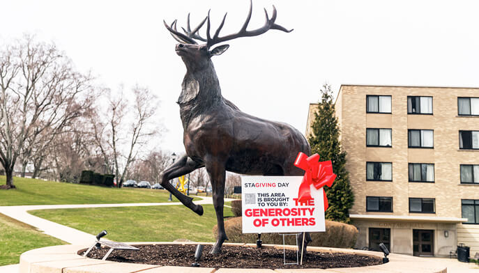A statue of a rearing stag with antlers on Fairfield University campus, a sign at its base reads, "This area is brought to you by the generosity of others," with a red ribbon.