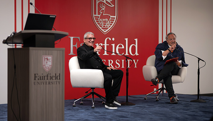 Stephen Wilkes and Philip I. Eliasoph sit on stage at Fairfield University, smiling and engaging with the audience. One holds a microphone. The atmosphere is lively and conversational.