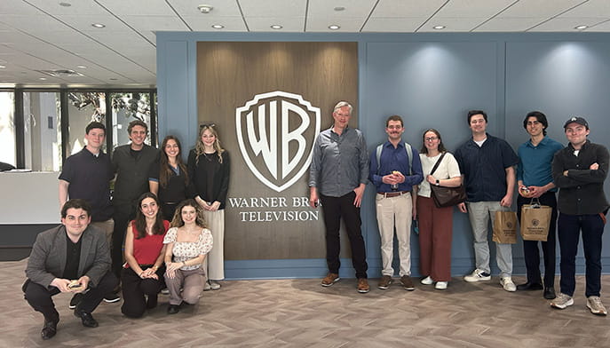 A group of Fairfield students, smiling and casually dressed, stand in a modern lobby with a large Warner Bros. Television logo on the wall behind them.