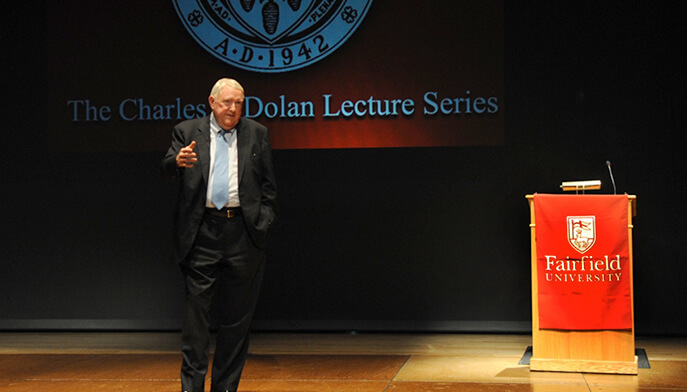 E. Gerald Corrigan ’63, PhD in a suit gestures passionately while speaking at a podium. Behind him, partially visible text reads "Lecture Series," indicating a formal setting.
