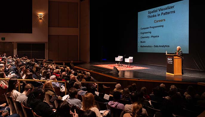 A speaker addresses a large crowd in an auditorium, engaging the audience with his presentation.