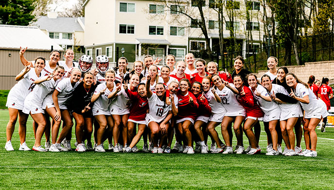 The women's lacrosse team stands on the field, united and smiling for a team photo.