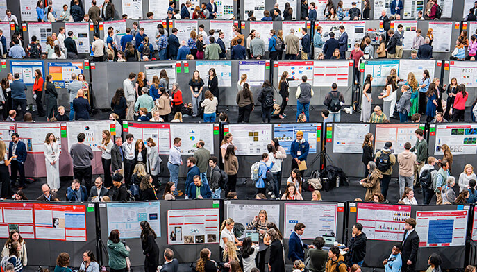 A crowded research symposium session with diverse groups engaging in discussions around various research posters. The atmosphere is active and collaborative.