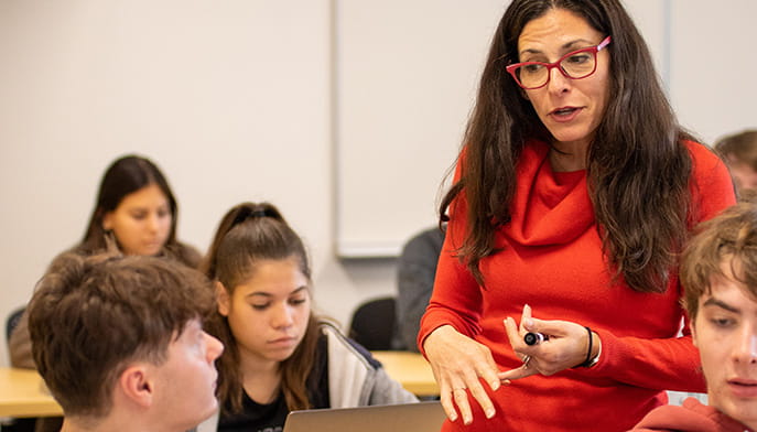 Professor Bloch in a red sweater engages with attentive students in a classroom. They sit with laptops open, creating a focused and interactive learning environment.
