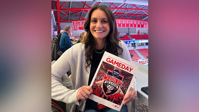Smiling Marissa Consiglio holds a "Gameday" program for the Mahoney Classic in an indoor sports arena, surrounded by red banners and people in the background.