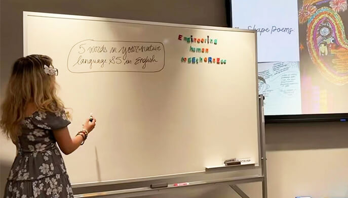A woman in a floral dress writes on a whiteboard. The board displays colorful letters and a text bubble.