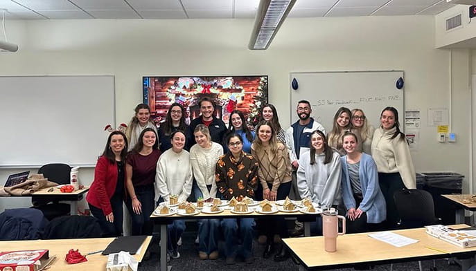 A group of smiling people stand in a classroom, gathered around a table with gingerbread houses. The room has a festive, holiday feel.