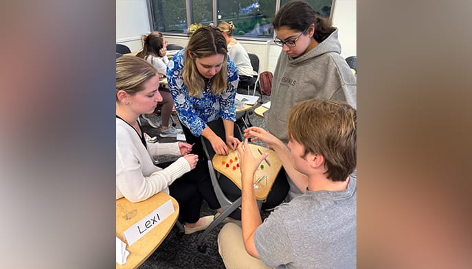 Four students collaborate in a classroom, playing and discussing an educational game with gummy candy and toothpicks. The scene feels focused and educational.