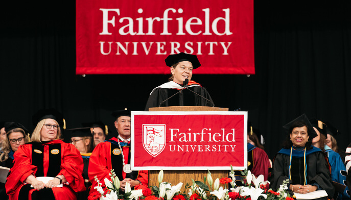 Maggie Doyne in academic regalia stands at a podium with a Fairfield University banner. Behind them, officials sit on stage, surrounded by flowers.
