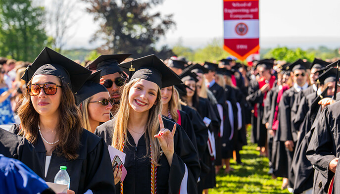 A joyful group of Fairfield graduates in caps and gowns stand outside on a sunny day. A woman in the front center smiles and gestures the “stags up” sign.
