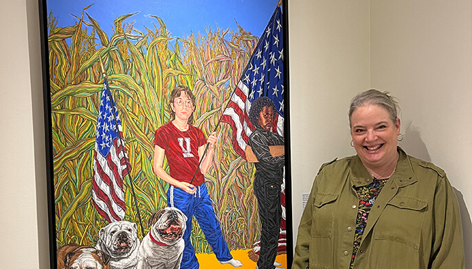 A smiling person, LeeAnn Weaver, stands beside the vibrant artwork Girl’s Army–the bitches by Katharine Kuharic, featuring a field of corn, two U.S. flags, and two bulldogs. The painting exudes patriotism and realism.