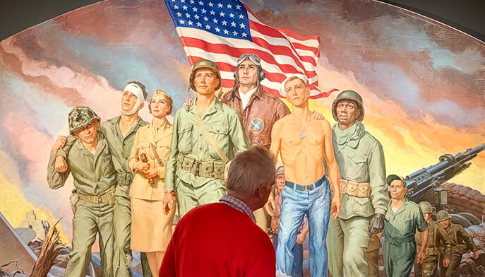 A person in a red sweater stands in front of a colorful mural, Robert Lynn Lambdin’s 1958 [Heroes of World War II] painting, in Meditz Gallery depicting diverse soldiers and a nurse, with an American flag in the background.