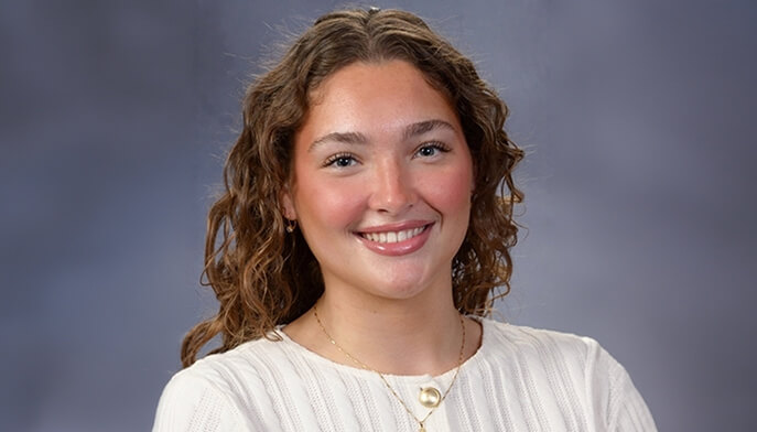 Caroline Cossman '26 with curly brown hair and a friendly smile poses in front of a gray background. She wears a white sweater, conveying a warm and approachable tone.