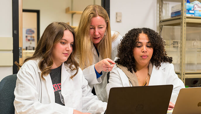 Three women in lab coats collaborate in a lab, focusing on laptops. The atmosphere feels studious and engaged, emphasizing teamwork and learning.