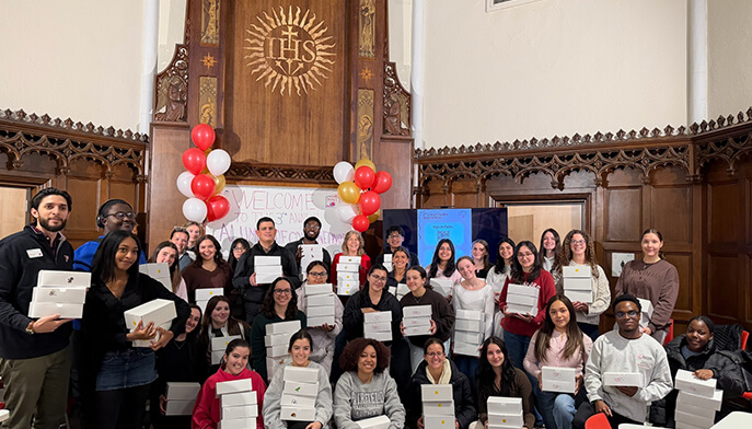 A diverse group of smiling people hold white boxes in a decorated room with red and white balloons and a wooden backdrop featuring a large IHS symbol.