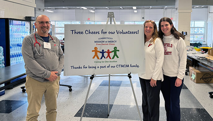 Three people stand smiling beside a sign at a volunteer event. The sign celebrates volunteers of the Connecticut Mission of Mercy dental clinic.