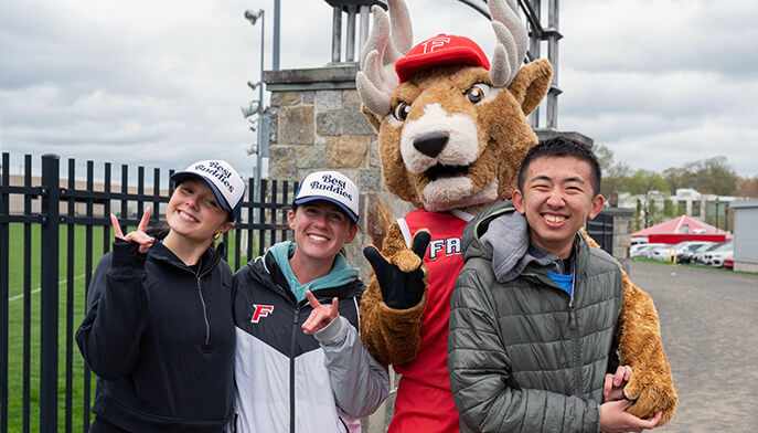 Three smiling students pose with Lucas the Stag mascot. They stand outdoors near a fence, exuding a cheerful and playful vibe.