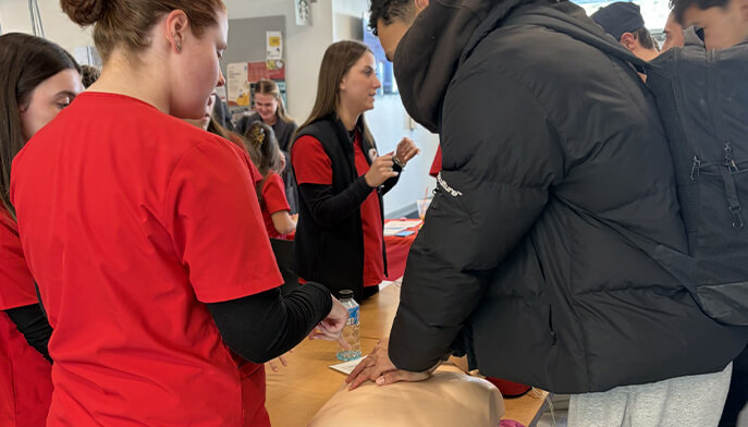 Students in red shirts supervise a CPR training session with a mannequin, participants attentively practicing chest compressions on a table.