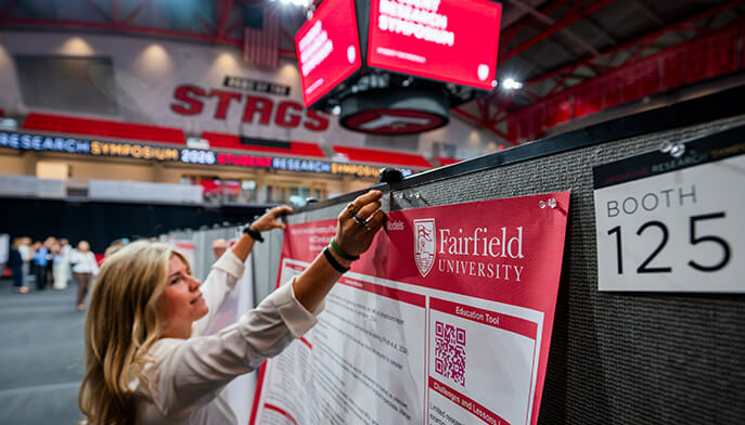 A student hangs a Fairfield University poster at a research symposium in an indoor venue. Booth 125 sign visible, with a professional, focused atmosphere.