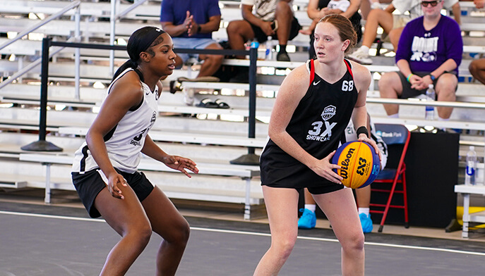 Two female basketball players in action on an outdoor court. One, in a black jersey, holds the ball while being closely guarded by a player in white. Spectators in the background watch intently.