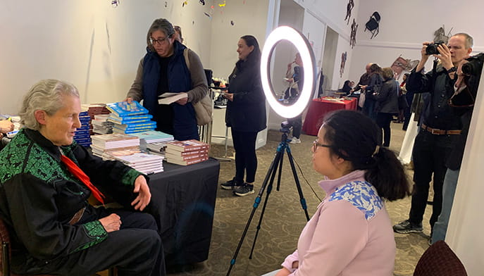 Two people are seated and talking in front of a ring light and stacks of books while others stand and a photographer takes pictures in an indoor event space.